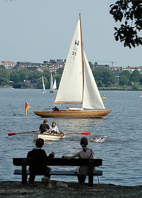 011_14469 - viel zu sehen auf der Alster; ein Segelschiff wird vorbeigesteuert; das Ruderboot kommt an Land und wird von zwei SchwimmerInnen begleitet - auf der Bank im Schatten sitzt ein Paar und beobachtet die Szene, der Hund schlft.