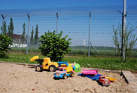 011_15853 - Sandkiste mit Kinderspielzeug - Bagger Lastwagen und Schaufel - im Gefngnis hinter dem hohen Metallzaun / Stacheldrahtzaun.