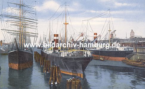 011_15708 - historische Hafenszene ca. 1900; ein Segelschiff und ein Frachter liegen an Duckdalben in der Mitte des Hafenbeckens. Ein anderes Frachtschiff liegt am Kai und wird mittels Krane entladen; dahinter sind Lagerschuppen zu erkennen. 
