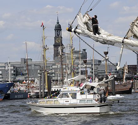 011_17502 - Bilder von Segelschiffen in Hamburg - yachthafen |ein Matrose refft im Bugspriet eines Grossseglers die Segel; davor f�hrt eine Motoryacht in den Hamburger Yachthafen am Baumwall. Hinter den Masten der Segler die Silhouette des Verlagsgeb�ude von Gruner + Jahr am Baumwall, dahinter die Hamburger St. Michaeliskirche.