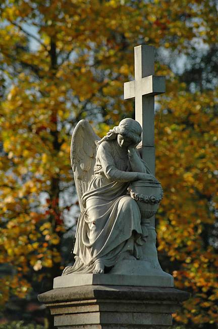 trauernder Friedhofsengel vor gelben Herbstlaub - der Engel kniet vor einem Kreuz und hat den Kopf in die Hand gesttzt. 
