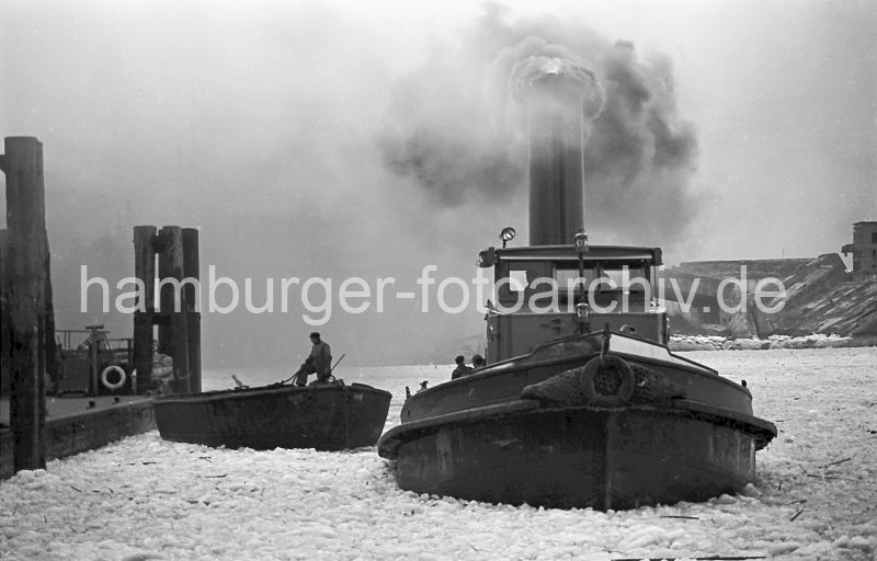 Winter im Hafen Hamburgs - Schlepper und Schute im Vulkanhafen; 1956 219_10_051 Ein Schlepper unter Dampf im Vulkanhafen - dick steigt der Qualm in den winterlichen Hamburger Himmel; das Wasser des Hamburger Hafens ist mit Treibeis bedeckt. Ein Schiffer auf einer Schute hat ein Tau in der Hand und bereitet sich darauf vor, das Schiff am Anleger festzumachen. Im Hintergrund Trmmer des U-Boot Bunkers Elbe 2; in dem 1941 fertig gestellten Bunker wurden die auf der Howaldtswerft gebauten U-Boote ausgerstet. Der Bunker hatte eine Deckenstrke von 3m - nach Kriegsende wurde die Anlage von britischen Truppen mit 47 Waggonladungen Fliegerbomben gesprengt.