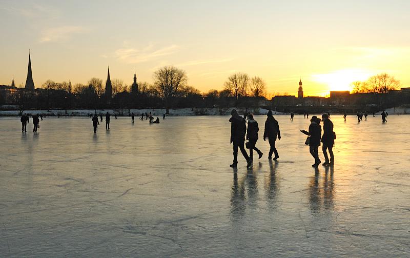 257_5875 Spaziergnger dem Eis der zugefrorenen Alster gehen Richtung Hamburg - St. Georg. Das Licht der untergehenden Sonne spiegelt sich auf der blanken Eisdecke