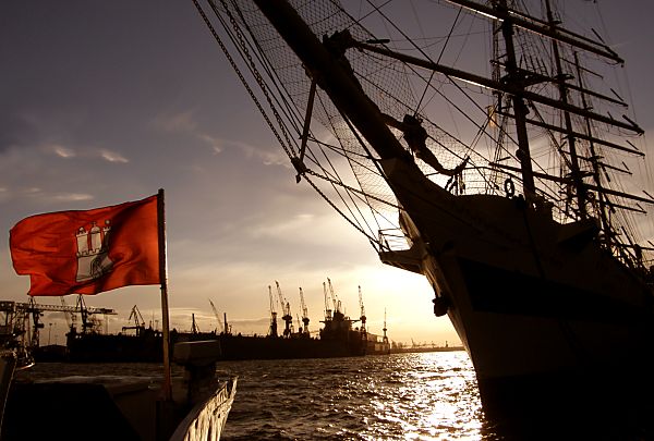 011_15708 - Sonnenuntergang im Hamburger Hafen; die Hamburg Fahne wird von der Sonne beschienen, vom Grosssegler sind Bug und Masten in der Silhouette zu erkennen - im Hintergrund Krne der Werft an der Elbe.