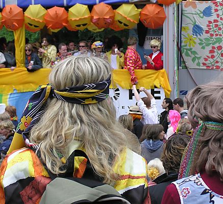 011_14214 - Mzwei als Hippie mit Langhaarpercke und Stirnband verkleidete Besucher auf dem SchlagerMove stehen am Strassenrand; auf dem Musikwagen oben recht ist ein blondes Heino - Imitat zu erkennen. 