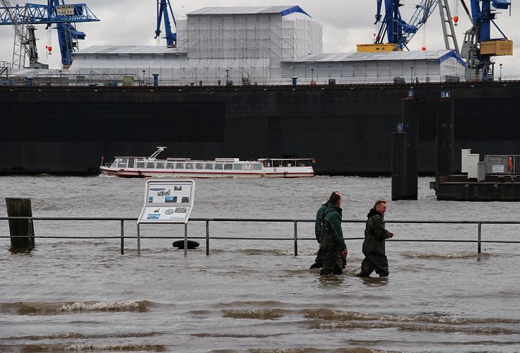 Hamburg Fotos Sturmflut - Elbhochwasser auf dem Hamburger Fischmarkt St. Pauli  33_47911 Die Sturmflut hat das Gelnde des Hamburger Fischmarkts unter Wasser gesetzt. Die Elbe ist ber die Kaimauer getreten und bersplt das Gelnde auf dem Sonntags die Stnde des Fischmarkt aufgebaut sind. Drei Katastophenhelfer waten durch das kniehohe Wasser; im Hintergrund das Trockendock der Hamburger Werft Blohm + Voss, davor fhrt eine Barkasse der Hamburger Hafenrundfahrt. www.fotos-hamburg.de