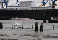 33_47911 Die Sturmflut hat das Gelnde des Hamburger Fischmarkts unter Wasser gesetzt. Die Elbe ist ber die Kaimauer getreten und bersplt das Gelnde auf dem Sonntags die Stnde des Fischmarkt aufgebaut sind. Drei Katastophenhelfer waten durch das kniehohe Wasser; im Hintergrund das Trockendock der Hamburger Werft Blohm + Voss, davor fhrt eine Barkasse der Hamburger Hafenrundfahrt. www.fotos-hamburg.de