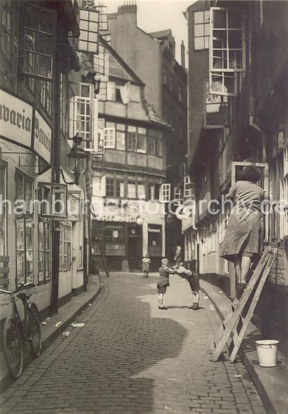 Historische Hamburger Architekturfotografien. Schmale Gasse mit Kopfsteinpflaster in einem Gngeviertel.  04_076_1 Als Gngeviertel wurden die besonders eng bebauten Wohnquartiere in einigen Teilen der Hamburger Altstadt und Neustadtbezeichnet. Die Gngeviertel waren grtenteils mit Fachwerkhusern bebaut, deren Wohnungen zumeist nur durch schmale Straen und ber Hinterhfe und den Gngen zwischen den Husern zu erreichen waren.  In den Gngevierteln wohnten meist mittlere und rmere Bevl- kerungs schichten; aufgrund der schlechten hygienischen Zustnde, aber auch aus sozialen und politischen Bestreb- ungen begannen bereits zum Ende des 19. Jahrhunderts die ersten Sanierungsmanahmen durch Abriss.