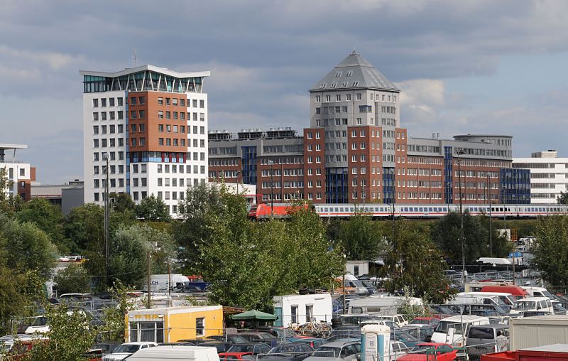 Fotografien aus den Hamburger Bezirken - Bezirk HH- Mitte, Stadtteil Rothenburgsort.Gebude der Behrde fr Stadtentwicklung und Umwelt - Gebrauchtwagenhandel. 471_ 4598 Moderne Architektur der Behrde fr Stadtentwicklung und Umwelt sowie das Amt fr Gesundheit und Verbraucherschutz. Auf den ehemaligen Gleisanlagen des Rangierbahnhofs Rothenburgsort haben sich viele Autohndler nieder gelassen. Die einzelnen Areale sind mit Bauzunen abgetrennt.  Ein Regionalzug mit roter Lokomotive fhrt auf der Trasse vor den Verwaltungsgebuden.