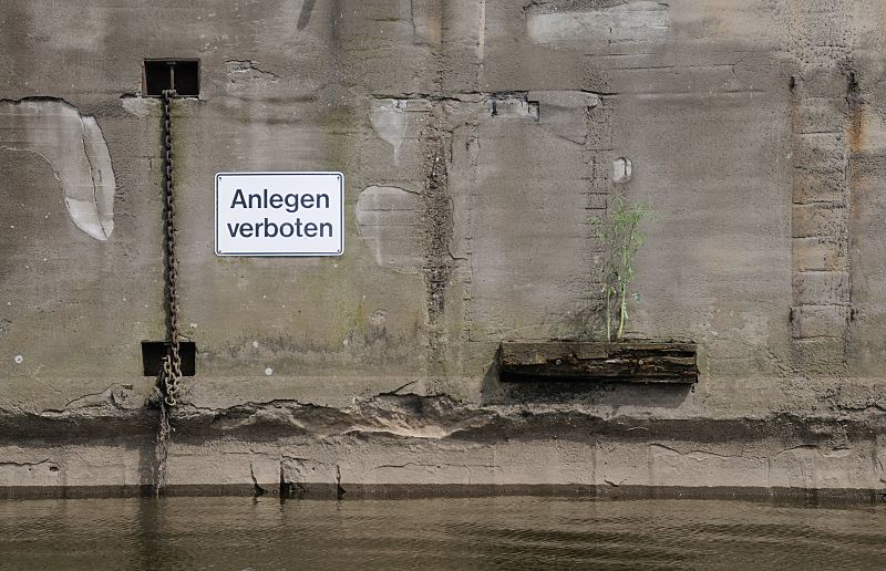 Bilder aus Hamburg - Fotos aus den Hamburger Stadtteilen: Peute / Veddel Kaimauer am Kanal - Anlegen verboten. Hamburg-Fotografie Christoph Bellin. 238_8417 EAn einer Kaimauer eines Veddeler Kanals ein weist ein Schild darauf hin, dass das Anlegen mit einem Schiff verboten ist. Eine Eisenkette hngt ins Wasser, auf einem Holzbalken wchst eine Pflanze.