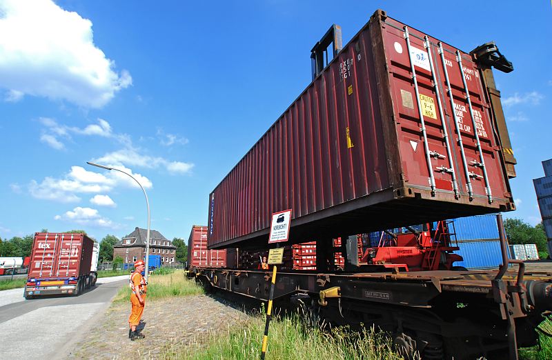 Containerverladung / Containerzug / Lastwagentransport  Fotos von der Arbeit in Hamburg Wilhelmsburg / Bezirk Hamburg Mitte 182_6946 Ein Container wird von einem Containercarrier auf den Gterwaggon eines Eisenbahnzugs geladen - ein Arbeiter in Signalkleidung und Helm berwacht die Arbeit. Auf der Strasse fhrt ein Sattelzug mit einem Containertransport.