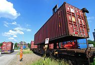 182_6946 Ein Container wird von einem Container Carrier auf den Güterwaggon eines Eisenbahnzugs geladen - ein Arbeiter in Signalkleidung und Helm überwacht die Arbeit. Auf der Strasse fährt ein Sattelzug mit einem Containertransport.