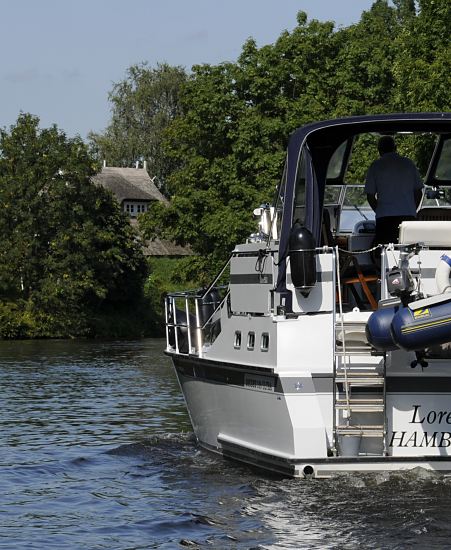 Hamburg Fotografie - Motorboot auf der Doveelbe  11_21582 Das weisse Motorboot fhrt in der Flussmitte der Dove- Elbe; der Freizeitkapitn steht am Steuerstand unter der Persenning als Schutz gegen die Sonne. Hinter dem Deich ist das Strohdach eines Wohnhauses zu erkennen. www.hamburg-fotograf.com