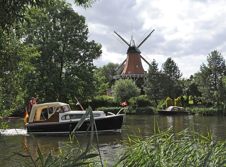 Fotos Sportbootrevier Hamburger Doveelbe - Reitbrooker Windmhle, Motorboot  11_21593 Die Dove-Elbe ist ein beliebtes Hamburger Gewsser fr Freizeitkapitne. Ein Motorboot fhrt flussabwrts, am Bug fhrt es die Hamburg-Fahne und am Heck die Deutschlandfahne. Die kleine Motoryacht passiert gerade die historische Reitbrooker Windmhle. Die Kornmhle wurde  ursprnglich 1773 errichtet und nach einem Brand 1870 neu erbaut. Am Ufer im Vordergrund wchst dichtes Schilf - auf der anderen Uferseite liegt am Bootsanleger ein kleines Sportboot.   www.hamburg-fotos.org