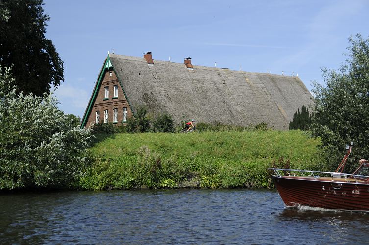 Hamburg Fotografie - Sommer in Bergedorf - Strohdachhaus am Deich, Motorboot 11_21598 Das Dach eines reetgedeckten Bauernhauses ragt hinter dem Deich der Doveelbe hervor. Die Deichstrasse ist in Hamburg eine sehr beliebte Strecke fr die bungsfahrten von Radrennfahrern. Eine Motoryacht fhrt flussabwrts auf der Dove-Elbe.  www.hamburg-fotos.org