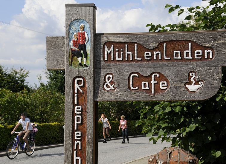 Hamburg Bilder, Bezirk Bergedorf - Schild Mhlenladen & Cafe Riepenburger Mhle   11_21637  Handgeschnitztes Hinweisschild zum Mhlenladen und Cafe bei der Riepenburger Mhle. Auf der Strassen fahren Inlineskater und ein Fahrrad. Die Strassen durch die Drfer der Vier- und Marschlande werden am Wochenende hufig von den Hamburger Ausflglern fr dieses sportliche Freizeitvergngen genutzt. www.hamburg-bilder.org