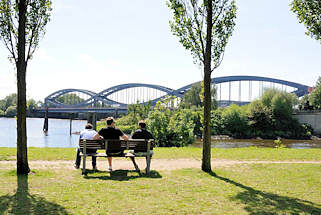 01178_5835 JParkbank in der Sonne an der Norderelbe im Elbpark Entenwerder - drei Parkbesucher sitzen auf der Bank und blicken aufs Wasser und die Norderelbbrcke.