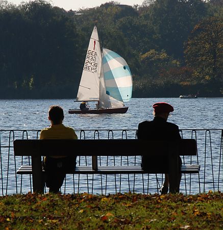 011_15810 - Herbstsonne am Alsterufer; zwei Spaziergnger ruhen sich auf einer Parkbank aus und beobachten ein Segelschiff im Sonnenlicht.  