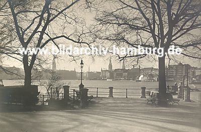 011_15921 - Ecke Lombardsbrcke / Neuer Jungfernstieg - Blick ber die Binnenalster; historische Fotografie ca. 1925 ) Im Hintergrund die Trme der Hansestadt Hamburg; im vorderen Bildteil eine Gaslaterne, Gelnder am Alsterufer und eine Bank am Baum. 