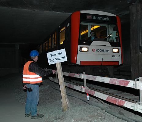011_14858 ein Zug der U2 Richtung Barmbek fhrt durch den Baustellenbereich; ein Schild Vorsicht Zug kommt weist auf die Gefahr hin; ein Arbeiter mit Helm und Signalhorn grt den Fahrer. 