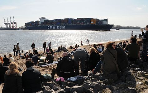 11_17450 - im Elbsand und auf Steinen sitzen die Gste in der Strandperle / Oevelgoenne in der Sonneund beobachten das Treiben auf der Elbe - der Containerfrachter wird mit Hilfe zweier Schlepper in den Waltershofer hafen geschleppt. 