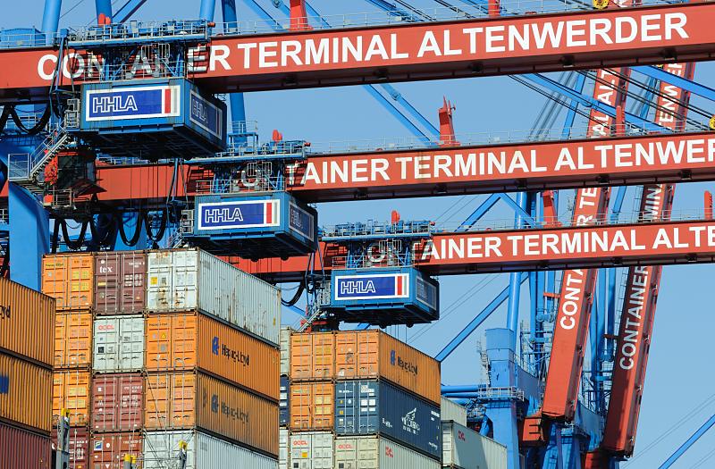 Bilder vom Containerhafen Hamburg - HHLA Container Terminal Altenwerder CTA. Containerbrcken und Laufkatzen ber der Schiffsladung eines Containerfrachters. 70_24610148 Drei der Hauptkatzen mit dem HHLA-Schriftzug befinden sich ber der Ladung eines Container- frachters um einen Container aus dem Laderaum zu Hieven und auf der Laschplattform abzustellen. Dort werden von Lascharbeitern die Twistlocks entfernt und die Metallbox von der Portalkatze zum Weitertransport auf eines der 75 AGV (Automated Guided Vehicle ) geladen.