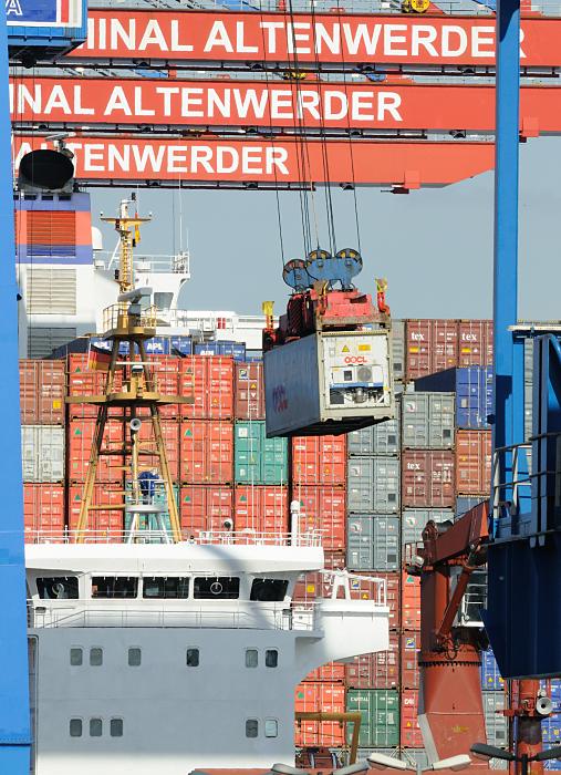Bilder vom Hamburger Containerhafen Entladung von Containern - Containerbrücken. 73_2433 Die Ladung eines Containerfrachters wird gelöscht - eine der genormten Metallboxen hängt hoch über dem Frachter an der Containerbrücke - im Hintergrund die gestapelten Container an Deck des Schiffs sowie dessen Aufbauten und Schornstein.