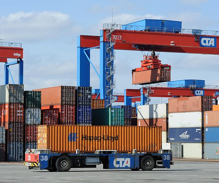 Fotos von der Arbeit im Hamburger Hafen - Container Terminal Altenwerder / CTA. Containertransport auf dem Betriebsgelnde Terminal Altenwerder. 83_1151 Einer der ca. 75 automatischen Transportfahreuge transportiert einen 40-Fuss (FEU) Container zum Portalkran - im Hintergrund hngt einer der Metallkisten hoch ber den gestapelten Container an dem Greifer eines Containerkrans.