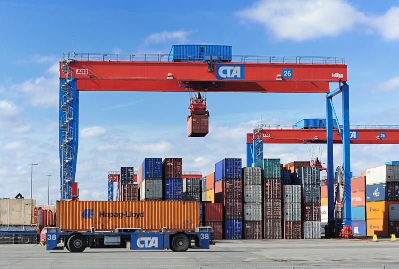 Bilddokumentation: Arbeit im Hamburger Hafen - Container Terminal Altenwerder / CTA. Containerverladung  auf dem Containerlager Terminal Altenwerder.  84_1155 Ein 40-Fuss (FEU) Standard-Container wird von den fahrerlosen Transportfahrzeug zu einem der Portalkrne gefahren. Einer der Container hngt am Kran und wird an seinen Lagerplatz auf einem der  26 Lagerblcken gebracht. .Jeder Block umfasst 10 Reihen  37 TEU;  an jedem Platz knnen vier - in den Auenreihen fnf - Container gestapelt werden. Die Krananlage besteht aus zwei unabhngig voneinander arbeitenden Krnen (Double Rail Mounted Gantry – DRMG), so dass gleichzeitig die Seeseite mit den AGV und die gegenberliegende Seite mit den Bahn und LKW bedient werden knnen. Durch ihre unterschiedliche Gre knnen beide Krne simultan ber den gesamten Block arbeiten, der kleinere Kran fhrt einfach unter dem greren durch.