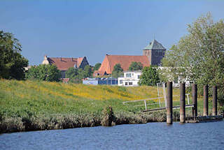 073_4791 Blick vom Flusslauf der Schwinge zur Stadt Stade; alte Eisenpoller am Ufer der Schwinge zeugen von dem ehemaligen Wirtschaftsverkehr, der auf der Fluss stattgefunden hat. Am Deich des Flusses blhen gelbe Blumen - im Hintergrund die dreischiffige gotische Hallenkirche St.