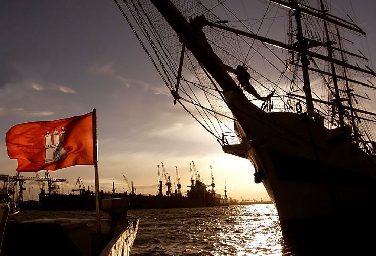 11_21357 Sonnenuntergang im Hamburger Hafen; ein Grosssegler liegt an den Landungsbrcken von Hamburg St. Pauli, die Segel sind gerefft. Der Bugspriet des Segelschiffs und Teilen der Masten sind als Silhouette am Hamburger Abendhimmel zu erkennen. Auf dem Bug einer Barkasse der Hamburger Hafenrundfahrt weht eine rote Hamburg Fahne im Wind. Im Hintergrund Krne und Dockanlagen der Werft Blohm & Voss. www.bildarchiv-hamburg.de