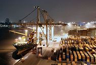 11_21439 Blick von der Containerbrcke am Athabaskakai; ein Containerschiff liegt an der Kaimauer und wird beladen. Starke Strahler beleuchten den Frachter und das Terminal Burchardkai - ein Container wird gerade von der Containerbrcke aufgenommen, um an Bord des Schiffes gebracht zu werden; Van-Carrier bringen neue Fracht vom Containerlager. Am anderen Elbufer die Lichter der Brogebude von Hamburg Neumhlen und auf der rechten Seite die blau angestrahlten Fautrme des Klrwerks Dradenau.  www.bildarchiv-hamburg.de