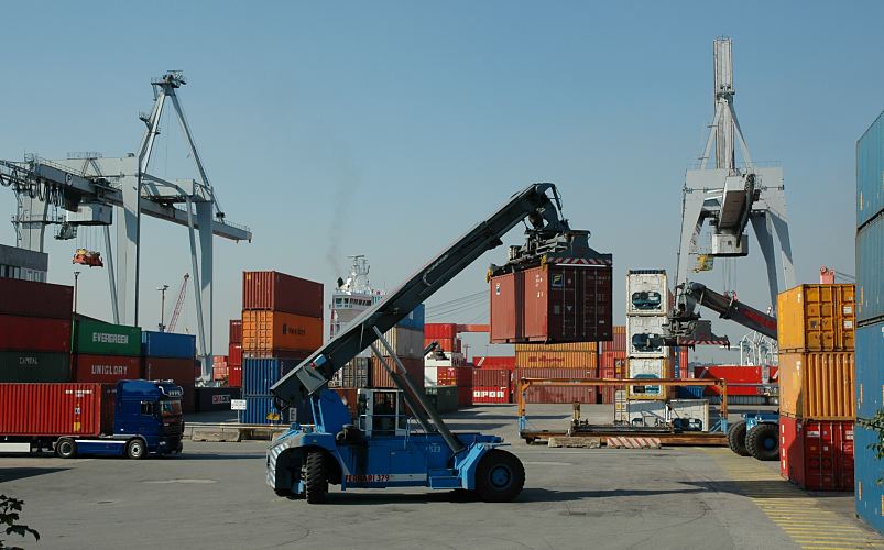 11_21442 Bilder vom Containertransport im Hamburger Hafen - Containerstapler und Lastwagen - Fotos vom Containerumschlag - Der Containerstapler hat einen Container von seinem Lagerplatz geholt und bringt seine Fracht zu Lastkraftwagen, die auf Tiefladern die Metallboxen weiter transportieren. Im Hintergrund liegt ein Containerschiff am Kai, seine Ladung wird ber Containerbrcken gelscht und neu befrachtet.   www.bildarchiv-hamburg.de