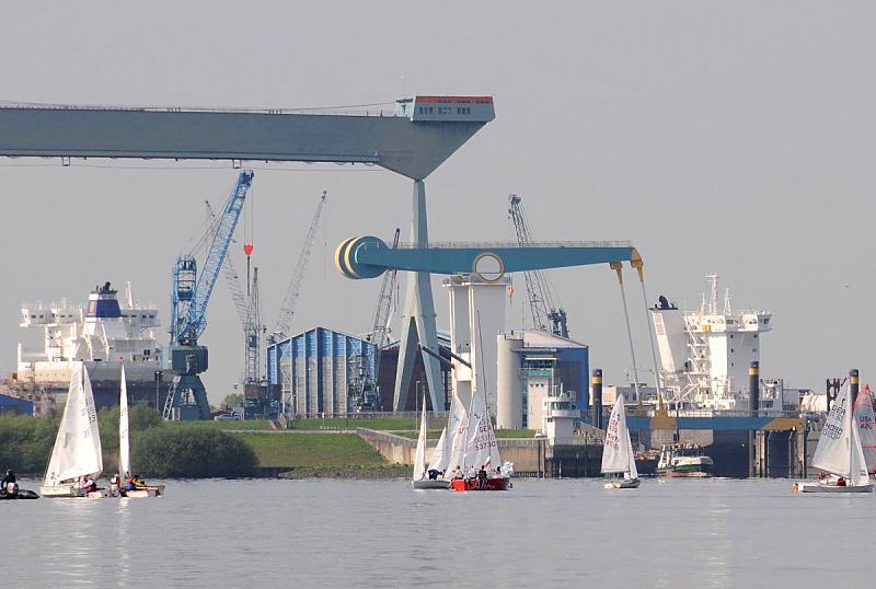 Hamburg-Bilder vom Hafen + der Elbe - Sietas Werft Hamburg Neuenfelde; Estesperrwerk. Fotoleinwand + Foto-Poster von Hamburgbildern als Geschenk zum Jahrestag - Jubilum - Firmenabschied. 31_8300 Die Elbe bei Neuenfelde - an der Klappbrcke und dem Este- Sperrwerk (re.) legt gerade die Fhre nach Hamburg Blankenese zum gegenberliegenden Elbufer ab - Jollen segeln auf der Elbe. Hinter dem Elbdeich liegt die Sietas Werft mit der gewaltigen Krananlage und kleineren Rollkrnen; lks. liegt ein Frachtschiff im Dock, whrend re. die Aufbauten eines Schiffs zu erkennen ist, das am Este-Ufer der Werft im Werfthafen liegt. 