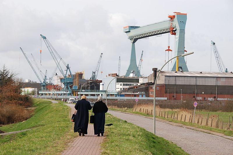 Fotos von der Hamburger Sietas Werft in Neuenfelde; Elbdeich + Werftanlage.. Fotoleinwand + Hamburg-Poster von Hamburgbildern als Geschenk zum Jahrestag - Jubilum - Firmenabschied. 33_8486 Am Neuenfelder Fhrdeich gehen zwei Frauen auf dem Winterdeich Richtung Neuenfelde. Auf der anderen Straenseite liegt die traditionsreiche Sietas Schiffswerft, die ihren Ursprung schon 1635 hat. Blick auf die Werftanlagen mit Krnen und Arbeitshallen, die sich an der Este-Mndung bei Hamburg Neuenfelde befinden. www.hamburg- bilder.org