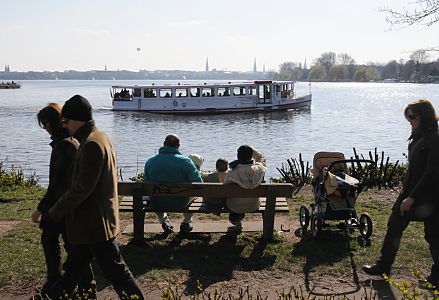 11_17424 |  Blick vom Alsterufer ber die Binnenalster; eine Familie mit Kindern sitzt auf der Parkbank in der Sonne - andere wandern am Alsterufer entlang. Ein Alsterschiff fhrt Hamburg Touristen ber die Alster Richtung Krugkoppelbrcke. 