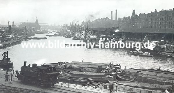 011_15733 - Foto vom historische Sandtorhafen ca. 1930; Frachtschiffe liegen am Sandtorkai und werden entladen  - die Fracht wird in den angrenzenden Lagerschuppen eingelagert. Im rechten Hintergrund die Backsteingebude der Hamburger Speicherstadt - die beiden Schornsteine gehren zum Kesselhaus.  Im Vordergrund liegen beladene Schuten am Kai - eine Lokomotive fhrt auf den Gleisen der Hafenbahn. Links im Hintergrund das Gebude vom Kaiserspeicher, dem Kaispeicher A. 