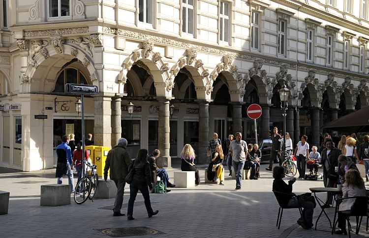 11_18977 Blick ber den Gustav-Mahler-Platz zum Sulengang der Hamburger Colonnaden. Auf dem Platz sitzen sitzen Gste in einem Strassencaf, andere haben sich in die Sonne gesetzt und essen dort Eis. Die Bgen ber den Sulen sind prchtig mit Stuckelementen dekoriert - ein Strassenschild weist darauf hin, dass die Colonnaden fr den normalen Durchgangsverkehr gesperrt sind.  www.bildarchiv-hamburg.de