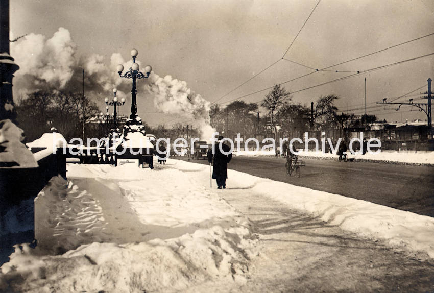 1085_X-0911-34 Es hat geschneit in Hamburg - hoher Schnee liegt auf der Lombardsbrücke; auf dem Bürgersteig ist ein Weg für die Fussgänger freigeschaufelt. Auf der Brücke über die Binnenalster fahren Kraftfahrzeuge, Fahrräder und ein Güterzug, dessen Lokomotive weissen Rauch in den Hamburger Himmel steigen lässt.