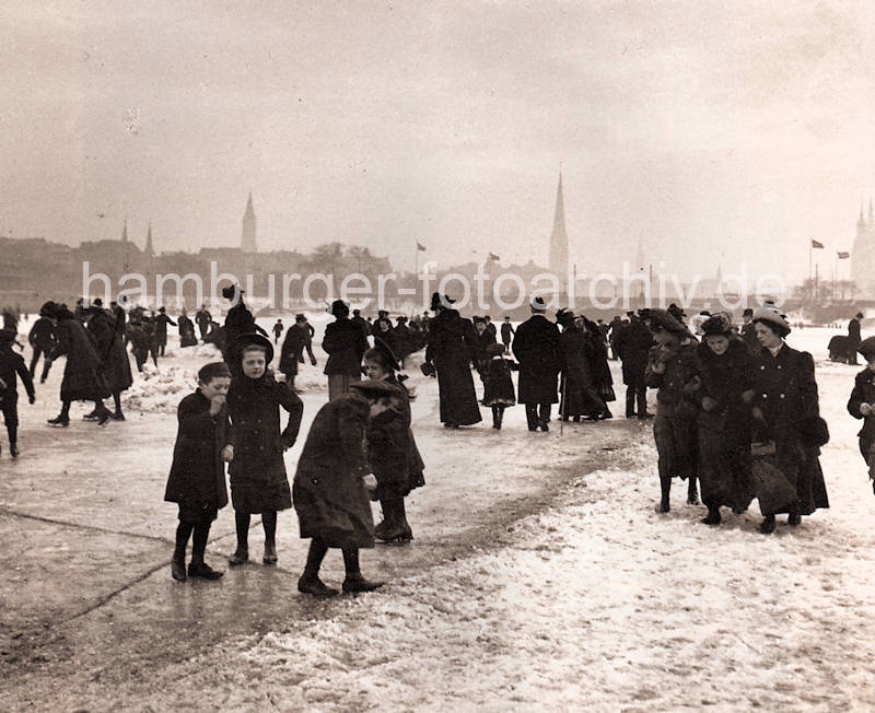 1086_3505_51 Kinder mit ihren Müttern auf dem Eis der zugefrorenen Aussenalster - einige haben Schlittschuhe untergeschnallt, andere glitschen auf dem Eis. Im Hintergrund Kirchtürme der Hansestadt Hamburg.