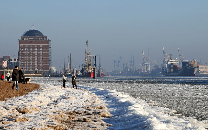 Fotos vom Hamburger Winter - Spaziergänger in der Wintersonne an der Elbe 11_22735 Spaziergänger geniessen die Wintersonne am Elbstrand in Hamburg Neumühlen; das Elbufer ist mit Eis bedeckt. Im Hintergrund das ehemalige Kühlhaus das zur Seniorenresidenz umgebaut wurde sowie der Museumshafen von Oevelgoenne. ©www.fotograf-hamburg.de