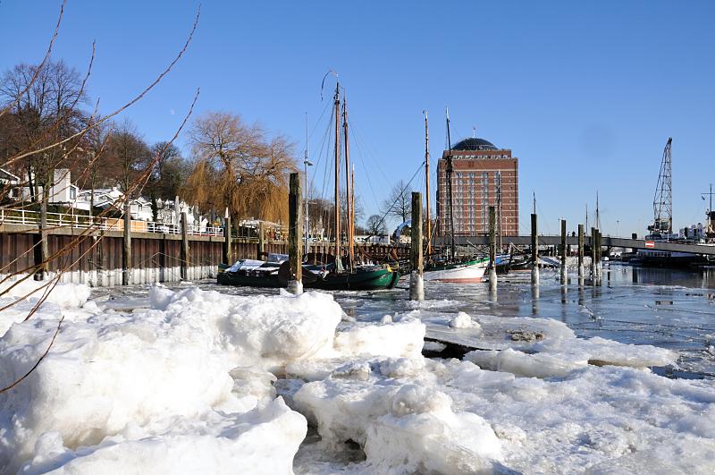 Hamburg Bilder im Winter - Museumshafen Oevelgoenne im Eis. 202_5723 Hoch aufgetürmt liegen die Eisschollen am Ufer des im Museumshafens im Hamburg Oevelgoenne. Einige der historischen Schiffe überwintern auf dem Wasser, andere lagern an einem Winterplatz. Hinter dem Steg zum Fähranleger der Hafenfähre das Gebäude des ehemaligen Kühlhauses in Neumühlen.