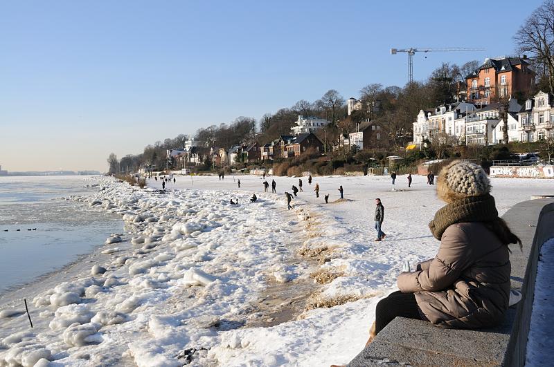 Fotograf von Hamburg - Hamburg-Motive vom Winter - Wintersonne am Elbufer. Spaziergaenger in der Sonne. 206_5730 Die Wintersonne scheint in Hamburg - Spaziergänger genießen das sonnige Wetter beim Spaziergang an der Elbe. Der Elbstrand ist mit Eis bedeckt, Kinder spielen am Ufer des Flusses. Am Hang stehen die Wohnhäuser von Hamburg Oevelgoenne.