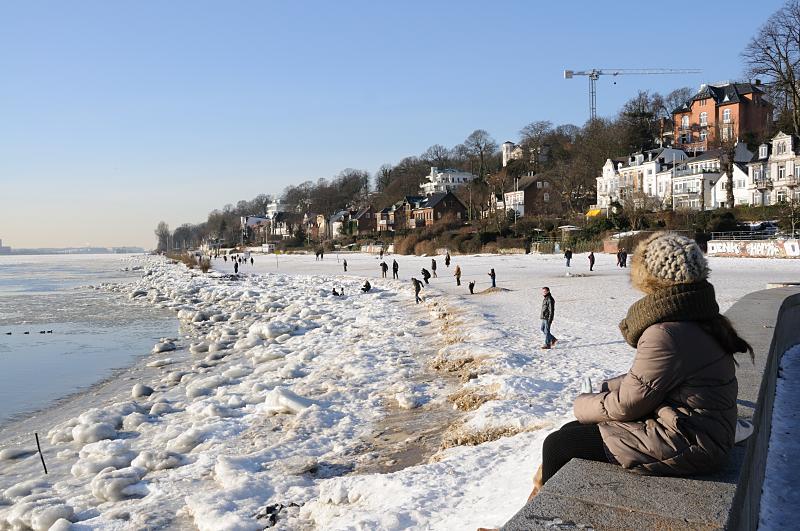 Fotograf von Hamburg - Hamburg-Motive vom Winter - Wintersonne am Elbufer. Spaziergaenger in der Sonne.  206_5730 Die Wintersonne scheint in Hamburg - Spaziergnger genieen das sonnige Wetter beim Spaziergang an der Elbe. Der Elbstrand ist mit Eis bedeckt, Kinder spielen am Ufer des Flusses. Am Hang stehen die Wohnhuser von Hamburg Oevelgoenne.