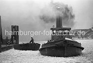 219_10_051 Ein Schlepper unter Dampf im Vulkanhafen - dick steigt der Qualm in den winterlichen Hamburger Himmel; das Wasser des Hamburger Hafens ist mit Treibeis bedeckt. Ein Schiffer auf einer Schute hat ein Tau in der Hand und bereitet sich darauf vor, das Schiff am Anleger festzumachen. Im Hintergrund Trümmer des U-Boot Bunkers Elbe 2; in dem 1941 fertig gestellten Bunker wurden die auf der Howaldtswerft gebauten U-Boote ausgerüstet. Der Bunker hatte eine Deckenstärke von 3m - nach Kriegsende wurde die Anlage von britischen Truppen mit 47 Waggonladungen Fliegerbomben gesprengt.