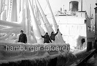 219_8_83 Die Mannschaft eines norwegischen Frachters, der gerade im Hamburger Hafen eingetroffen ist, steht an Deck des stark mit Eis bedeckten Schiffs. Die Decksaufbauten und Takelage des Frachtschiffs sind mit einer sehr dicken Eisschicht überzogen. 