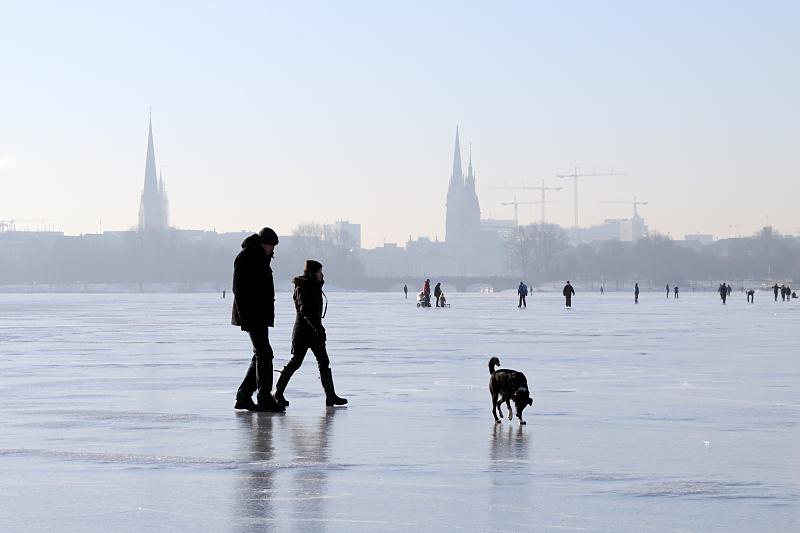 Jahreszeit Winter - Spaziergnger mit Hund auf der zugefrorenen Alster. Hamburger Fotograf - Auftragsfotografie, Pressefotografie || Schlittschuhlaufen mit Kinderwagen. 240_5501 Hamburger Spaziergnger nutzen den Sonnenschein und gehen mit ihrem Hund ber das blanke Eis der zugefrorenen Alster. Im Hintergrund die Kennedybrcke und die Trm der St. Petrikirchen und St. Nikolaikirche sowie der Turm des Hamburger Rathauses. 