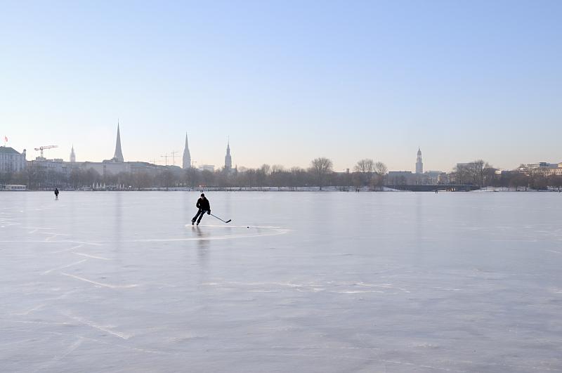 Hamburger Winter - frostiges Winterwetter, Sonnenschein. Hamburger Fotografie.  242_5558 Ein Eishockeyspieler dreht einsam seine Kurven auf dem Alstereis. Im Hintergrund das Panorama von Hamburg mit den Türmen der Hansestadt.