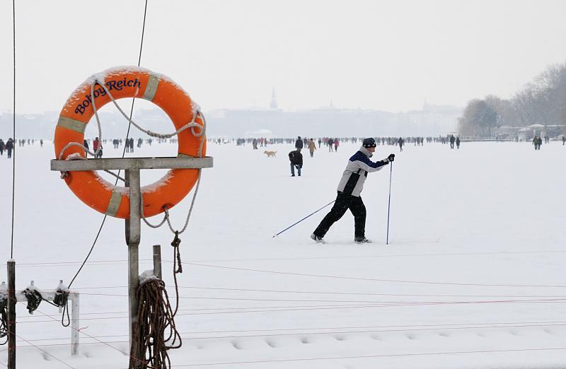 244_6093 Vor dem Anleger des Bootssteg vom Alstercaf Bobby Reich luft ein Hamburger Ski auf der zugefrorenen und verschneiten Alster; ein verschneiter Rettungsring hngt am Steg in seiner Halterung.  Hamburger Winter - Skilaufen auf der Aussenalster.