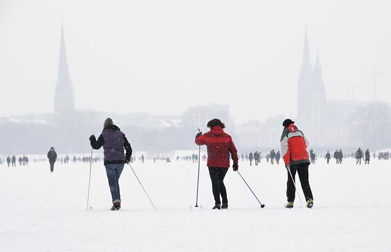 Hamburg im Winter - Schilanglauf auf dem Schnee der vereisten Alster. Verschneites + vereistes Hamburg. 245_6109 Skilangluferinnen nutzen den hohen Schnee, der auf dem Alstereis der Aussenalster liegt, um eine Runde um den 164 ha groen Hamburger Binnensee zu machen; die Rundstrecke am Alsterufer ist fr Jogger ca. 7,5 km lang. Im Hintergrund in den Schneewolken die Kirchtrme der St. Petri und Nikolaikirche sowie rechts der Rathausturm.  